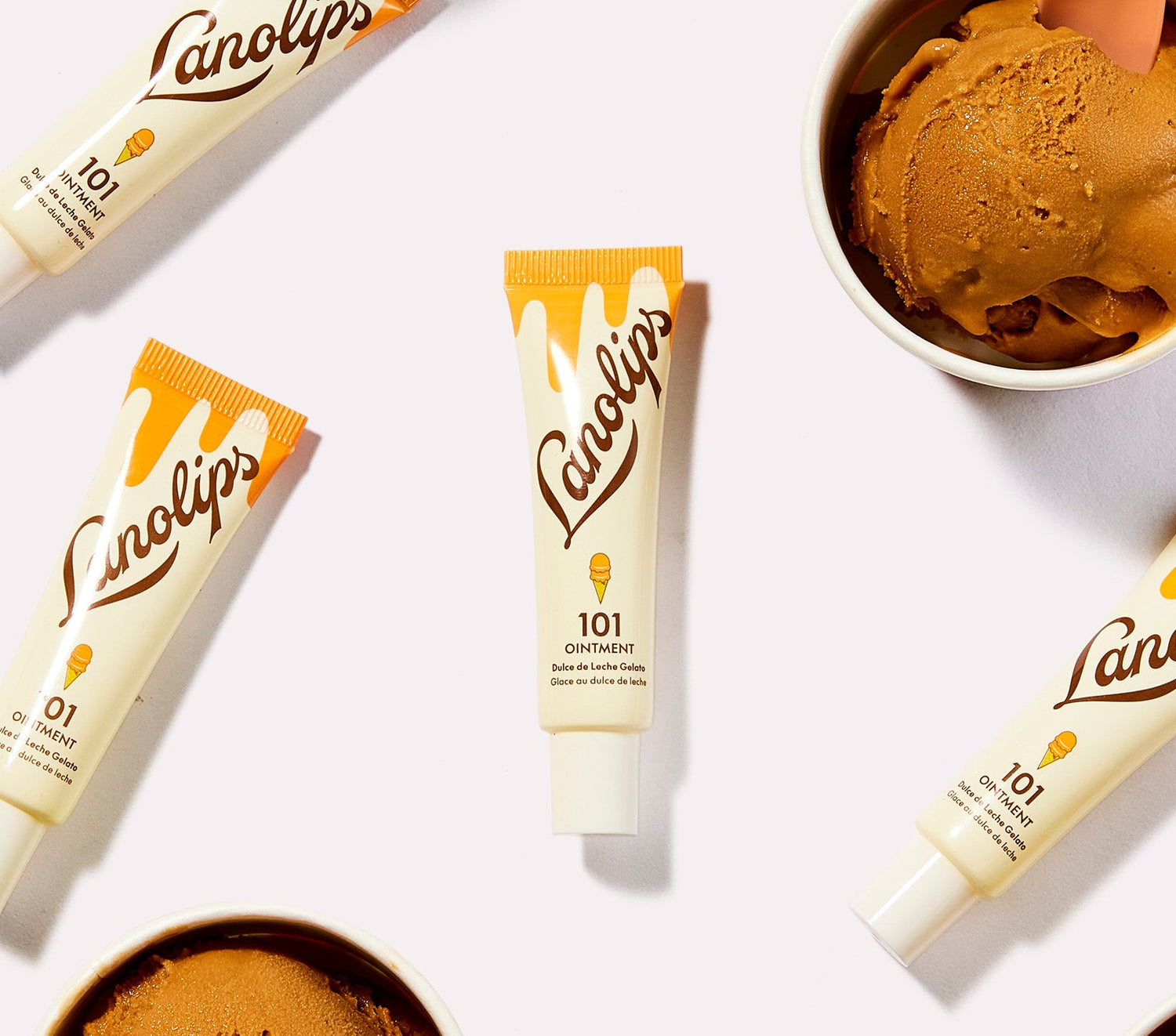 A flat lay image of multiple tubes of Lanolips 101 Ointment Multi Balm in Dulce de Leche Gelato. The tubes have a creamy, golden gradient with an ice cream cone illustration, complimenting the scoops of caramel-coloured ice cream placed nearby. The ointment is made from Aussie lanolin, with a hint of caramel designed to deeply hydrate dry lips.