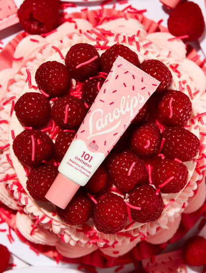 Flat lay of Lanolips 101 Ointment Multi-Balm in Raspberry Shortcake, placed on a swirl of pink frosting, fresh raspberries and sprinkles. This all-natural balm blends juicy raspberries with buttery shortcake for sweet hydration and a nourishing moisture boost.