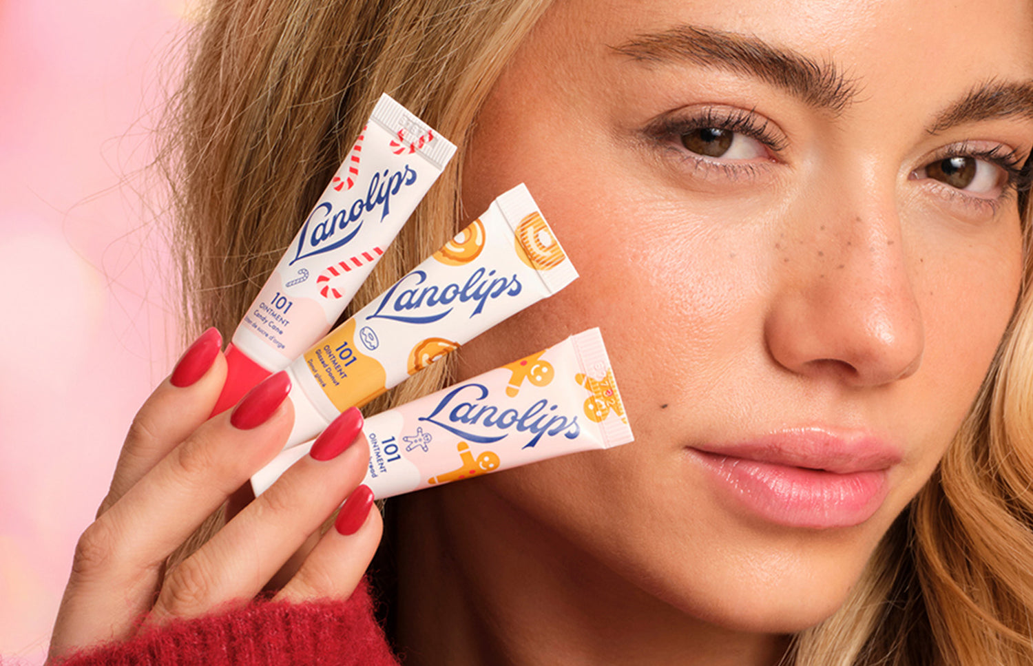 Model holding mini Lanolips lip balm tubes Candy Cane, Glazed Donut and Gingerbread in front of her face against a pink background. The minis are part of the  Lanolips Holiday Mini Trio. 
